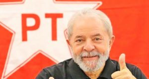 Lula fazendo joinha com o símbolo do PT atrás