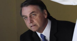 Foto de Bolsonaro com olhar de suspense, esperando algo. Ele usa terno preto com gravata azul.