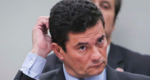 Foto de Sérgio Moro usando terno e com a mão direita levantada atrás da cabeça. Ele tem olhar preocupado.