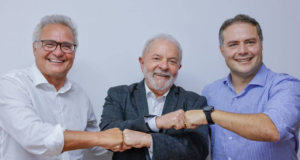 Foto de Renan Calheiros, (à esquerda), com camisa branca, óculos e cabelo branco. Lula ao centro com terno preto, barba e cabelo brancos. Renan Filho à direita, com camisa azul, sem barba e cabelo grisalho.