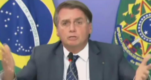 Bolsonaro surta e ataca esquerda. Foto do presidente usando terno cinza, fala ao microfone e tem os braços formando ângulo de 90°