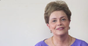 Dilma posando para foto, com camisa roxa e fundo branco. Ela sorri.