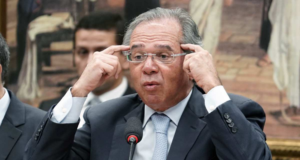 Paulo Guedes apontando para a própria cabeça, como se estivesse pensando. Ele está sentado, de óculos, terno e gravata e com um microfone à sua frente.