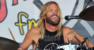 Imagem de Taylor Hawkins, de cabelo longo e louro, tocando bateria e sorrindo durante apresentação.