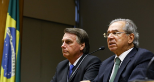 Foto de Bolsonaro ao lado de Paulo Guedes, ambos usam terno e estão sentados.