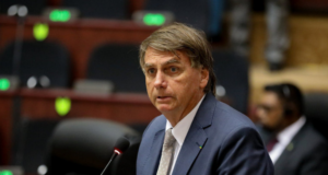 Foto de Bolsonaro com cara de susto. Ele fala ao microfone, usa terno azul e gravata cinza.