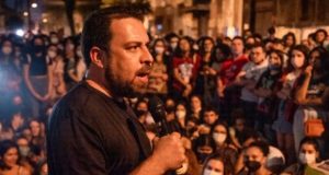 Guilherme Boulos de camiseta preta discursando em público com microfone na mão