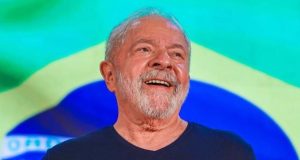 Luiz Inácio Lula da Silva de camiseta azul marinho, sorrindo, com a bandeira do Brasil ao fundo