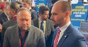 Alex Jones e Eduardo Bolsonaro lado a lado durante entrevista, os dois de terno, com outras pessoas próximas