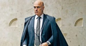 Alexandre de Moraes em pé, vestindo roupa azul marinho e com expressão séria