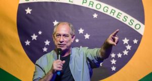 Ciro Gomes de camiseta preta e camisa social clara falando em microfone e apontando, com bandeira do Brasil ao fundo