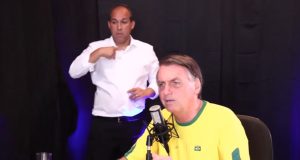 Jair Bolsonaro vestindo blusa do Brasil e falando em microfone com intérprete de libras ao fundo