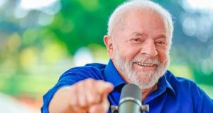 Lula de camisa social azul, sorrindo e apontando pra frente