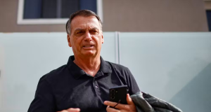 Jair Bolsonaro com camisa polo escura, falando e pedindo calma, carregando celular, óculos e casaco