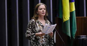 Gleisi Hoffmann falando em microfone com expressão séria, segurando papel, com roupa estampada