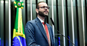 Jorge Seif de óculos, falando em microfone com bandeira do Brasil ao fundo