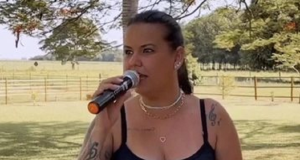 Nanda Ferraz cantando em microfone, de camiseta preta e rabo de cavalo, olhando para o lado