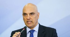 Alexandre de Moraes em fundo azul, falando em microfone, de terno e gravata, com expressão séria