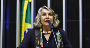 Érika Kokay falando em microfone, de roupa preta e lenço estampado, de cabelo solto