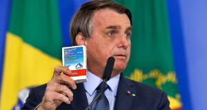 Jair Bolsonaro segurando caixa de cloroquina e olhando para o lado sério, perto de microfone e de bandeiras