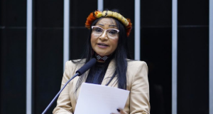 Silvia Waiãpi de roupa bege e arco de flores na cabeça, de óculos e cabelo solto, falando em microfone e segurando papel