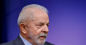 Lula de perfil, sério, de roupa social, em fundo roxo