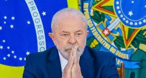 Lula sério, em posição de oração, com bandeiras ao fundo