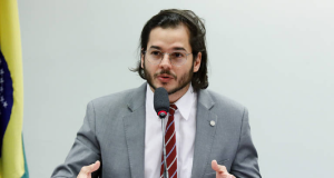 O deputado federal Túlio Gadêlha (Rede-PE) falando e gesticulando, de roupa social cinza e óculos, sem olhar para a câmera