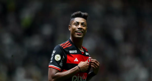 Bruno Henrique, atacante do Flamengo, segurando na camisa do time e sorrindo, em campo